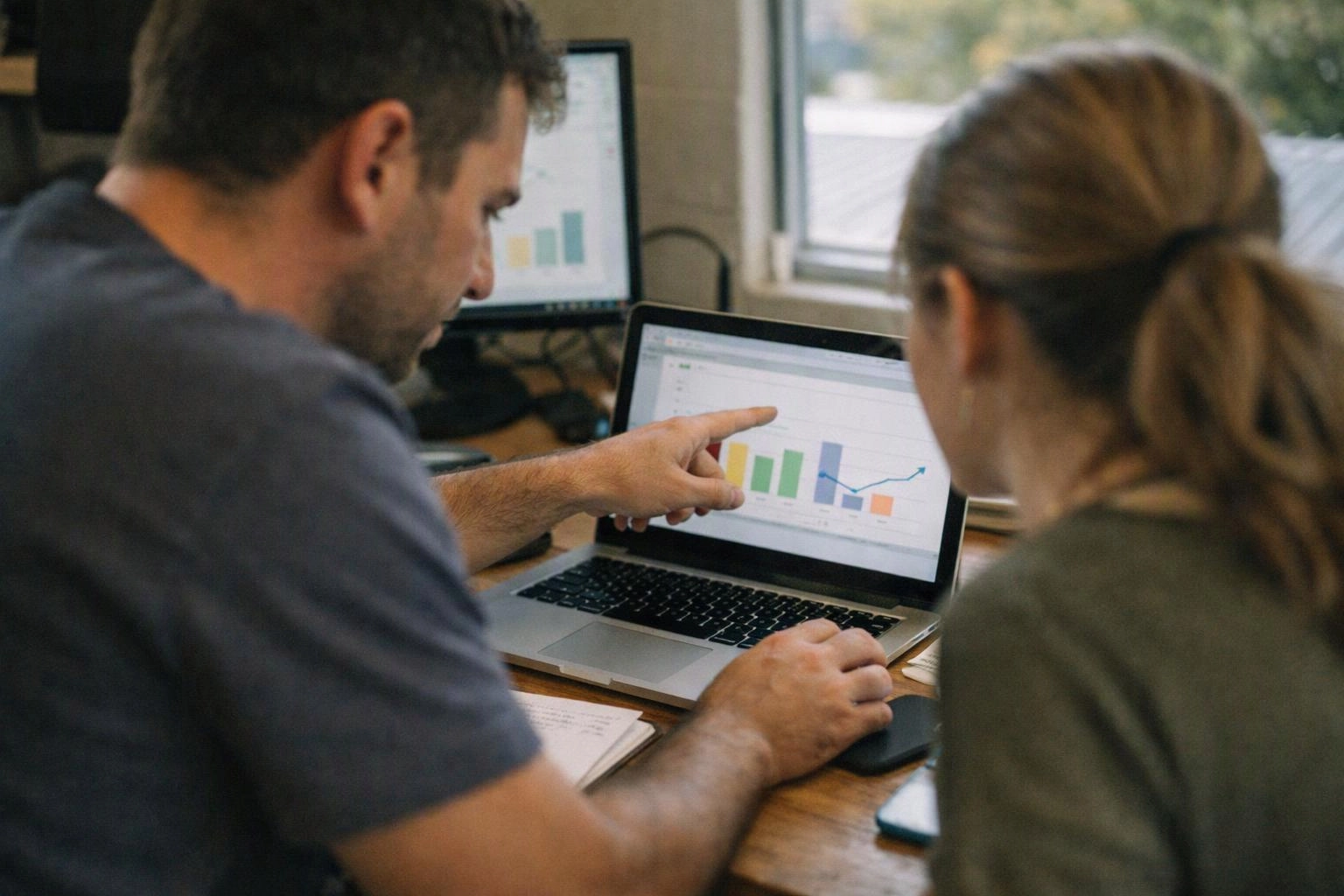 Two people sit at a desk, pointing at a laptop chart in a bright office