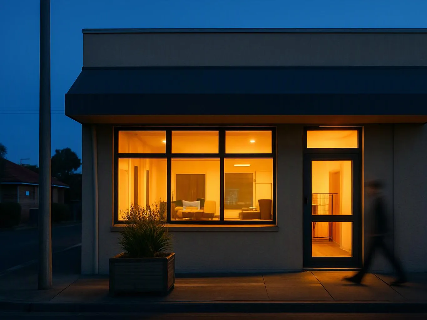 A lit storefront-like building stands at dusk, with warm windows, a potted plant, and a blurred person walking past the door