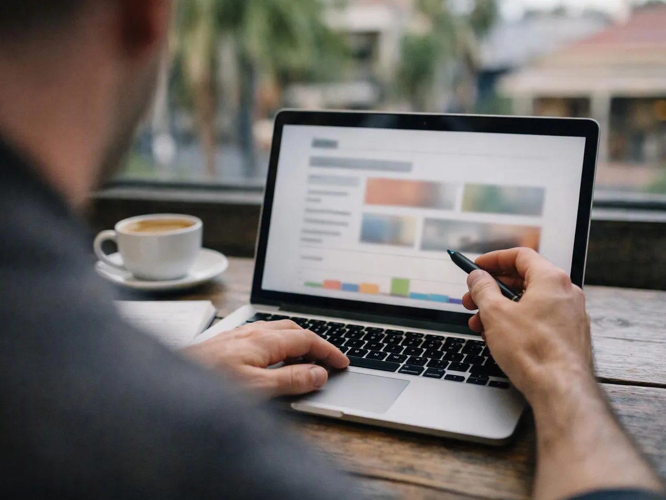 A person holds a pen over a laptop showing a blurred webpage, with a coffee cup beside it