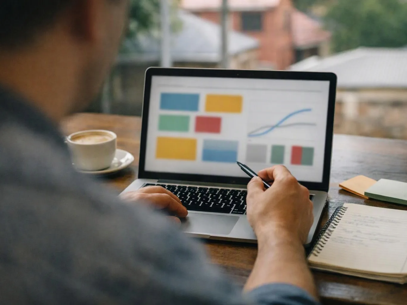 A person points at a laptop showing colorful charts beside a coffee cup and notebook on a wooden table