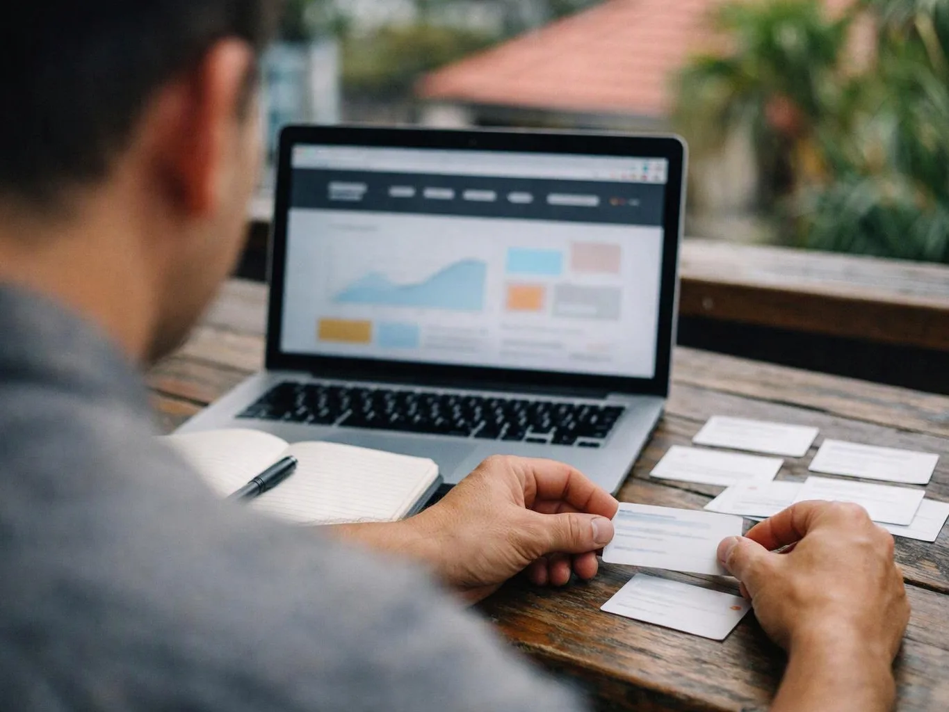 A person studies business cards beside a laptop with charts on a wooden table outdoors