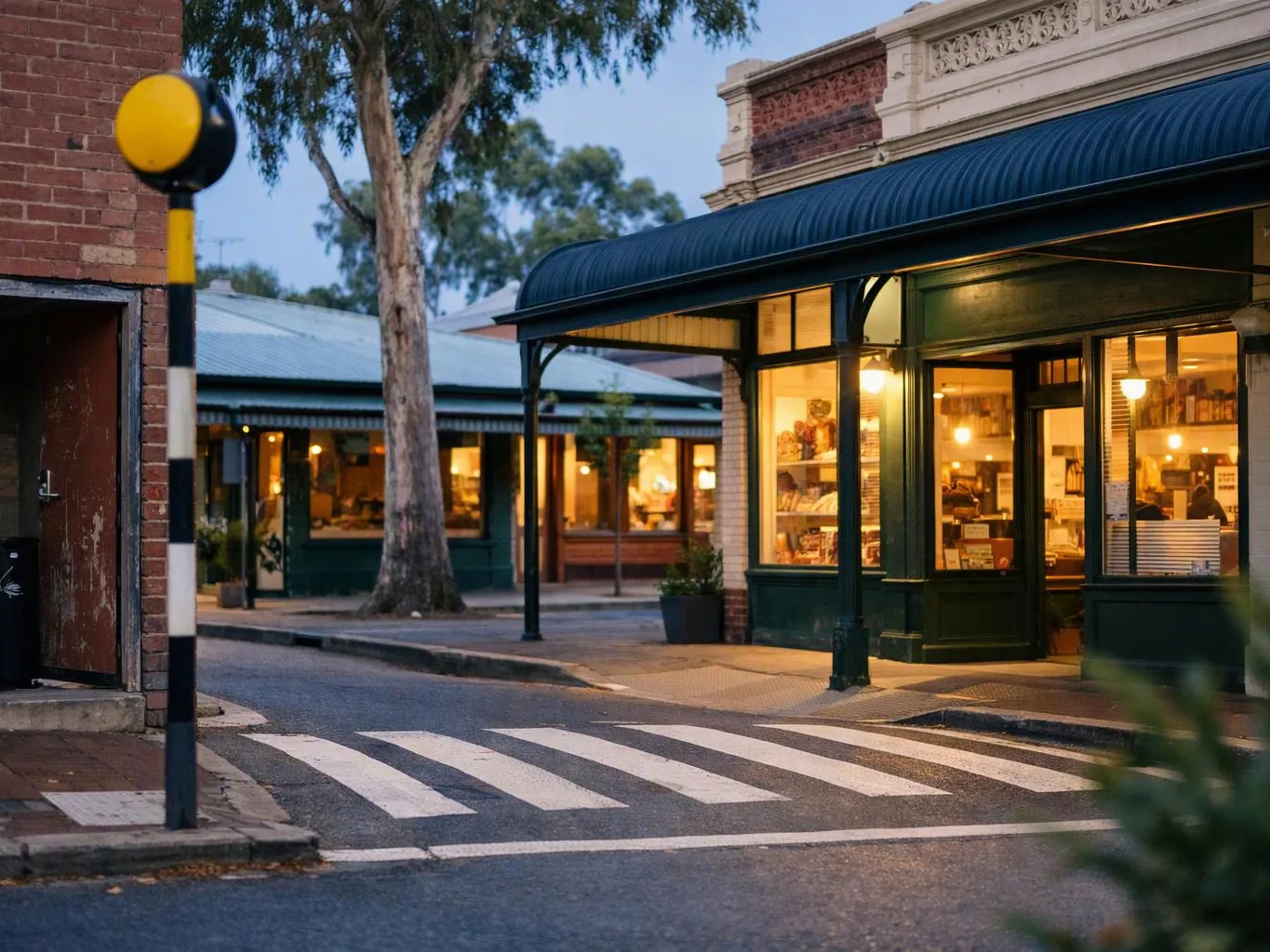 A quiet street corner shows lit storefronts, a zebra crossing, and a pedestrian signal at dusk