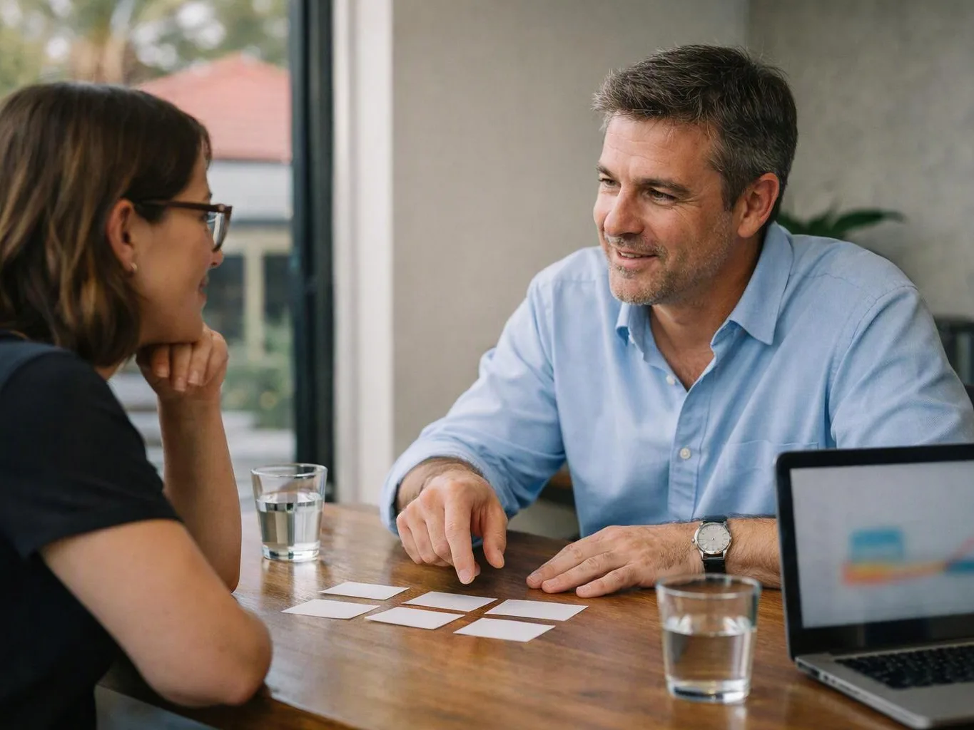 Two people sit at a table, talking over blank cards beside glasses of water and a laptop