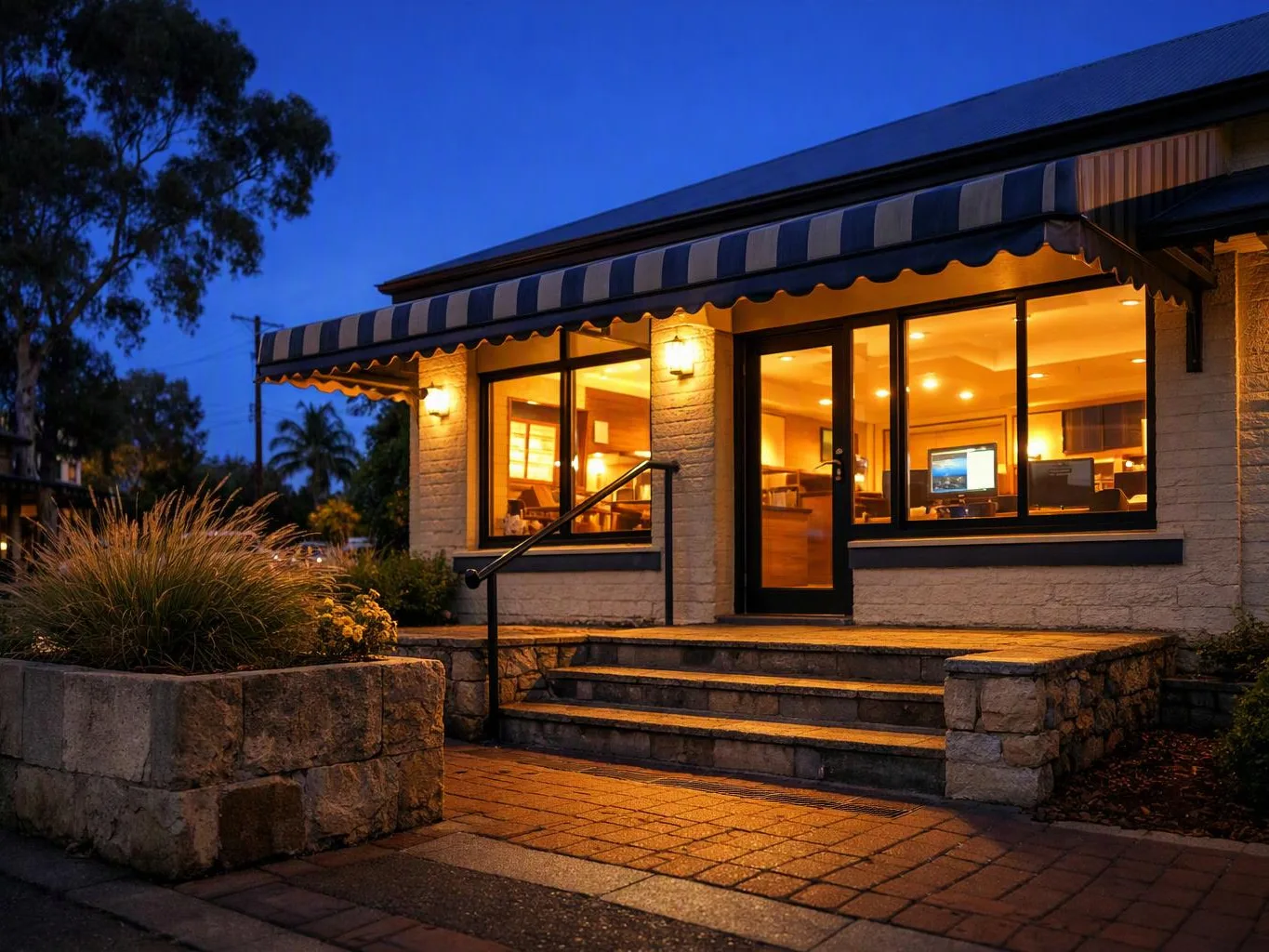 A warmly lit building entrance with striped awning, stone steps, and large windows at dusk